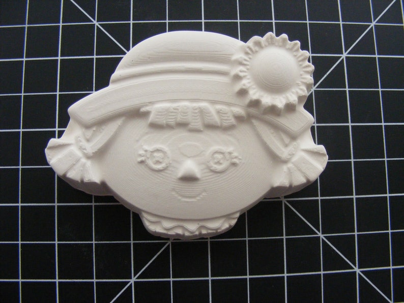 May include: White ceramic figurine of a girl with a hat and a flower. The girl has a simple face with eyes, a nose, and a smile. The figurine is a blank canvas for painting.