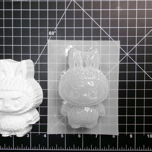 Puede incluir: Una figura de resina blanca con forma de conejo y un molde de silicona transparente. El conejo tiene una expresi&oacute;n severa y mide aproximadamente 7.6 cm de altura. El molde tiene la forma del conejo, listo para el moldeado de resina. El fondo es una cuadr&iacute;cula negra.