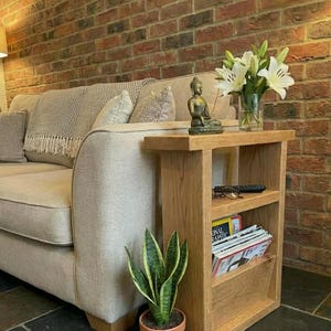 May include: A light oak side table with two shelves and a magazine rack, beside a beige sofa. The table holds a small Buddha statue, a vase of white lilies, and a remote control. A potted snake plant sits on the floor.