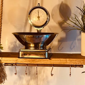May include: A classic kitchen scale with a circular dial and a stainless steel bowl. The scale is on a wooden shelf with a copper rod and hooks. A white pot with greenery hangs from the ceiling.