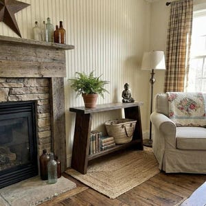 May include: A cosy living room with a wooden console table holding a potted fern, books, and a woven basket. A stone fireplace, a metal star, and a beige armchair create a warm, inviting atmosphere.
