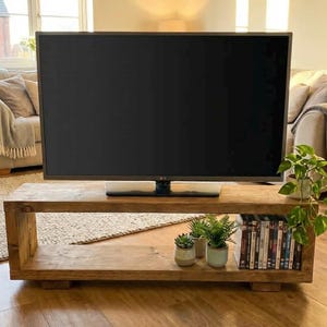 May include: A large flat-screen TV is placed on a rectangular wooden coffee table. The table has a lower shelf with DVDs and potted plants. The room has a neutral colour palette with natural light.