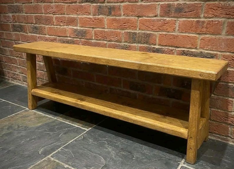 K&ouml;nnte beinhalten: Eine rechteckige Holzbank mit einer glatten, hellbraunen Oberfl&auml;che. Die Bank verf&uuml;gt &uuml;ber eine flache Sitzfl&auml;che und eine untere Ablage zwischen vier stabilen, geraden Beinen. Die Holzmaserung ist auf der gesamten Struktur der Bank sichtbar.