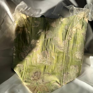 May include: A green corset with a floral pattern. The corset has a structured design with vertical seams and a sweetheart neckline. The fabric has a textured appearance and is adorned with delicate, sheer, white straps.