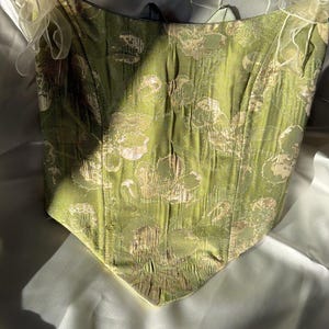 May include: A green corset with a gold and cream floral design. The corset features a sweetheart neckline and cream-coloured, sheer ribbon straps. The fabric has a textured finish, and the garment is set against a soft, white backdrop.