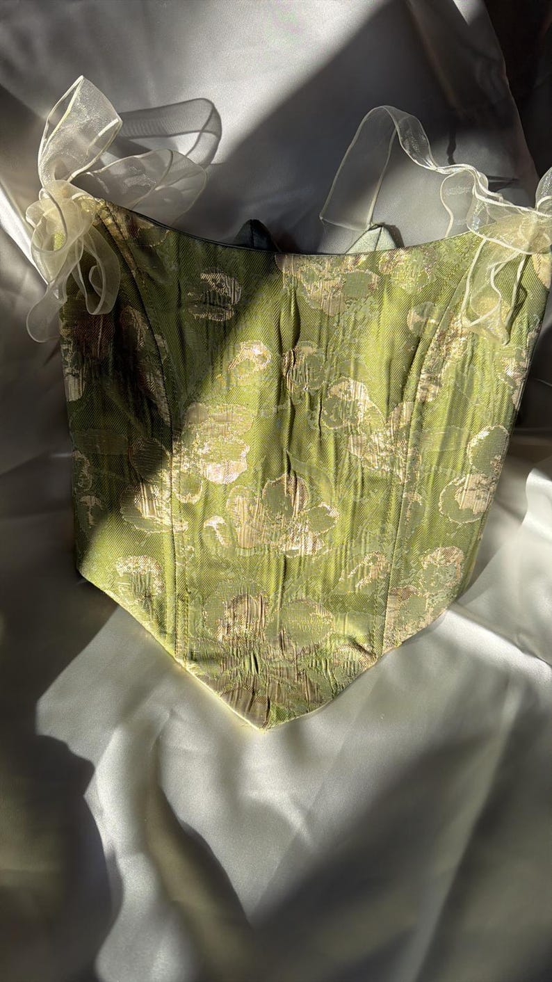 May include: A green corset with a gold and cream floral design. The corset features a sweetheart neckline and cream-coloured, sheer ribbon straps. The fabric has a textured finish, and the garment is set against a soft, white backdrop.