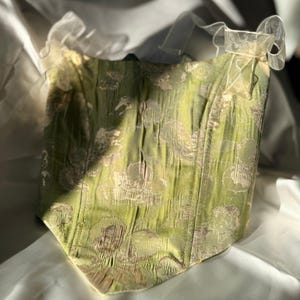 May include: A green corset with a floral design and a sweetheart neckline. The corset features a textured fabric and is embellished with delicate, sheer bows at the top. The garment is displayed on a white surface, with sunlight creating shadows.