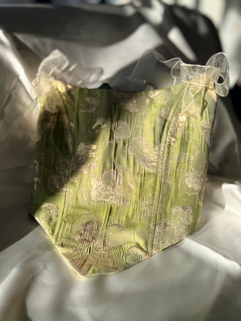 May include: A green corset with a floral design and a sweetheart neckline. The corset features a textured fabric and is embellished with delicate, sheer bows at the top. The garment is displayed on a white surface, with sunlight creating shadows.