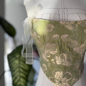 May include: A green and gold floral patterned corset top with a sweetheart neckline. The corset is displayed on a white dress form and features a delicate, sheer, white ribbon detail at the shoulder.