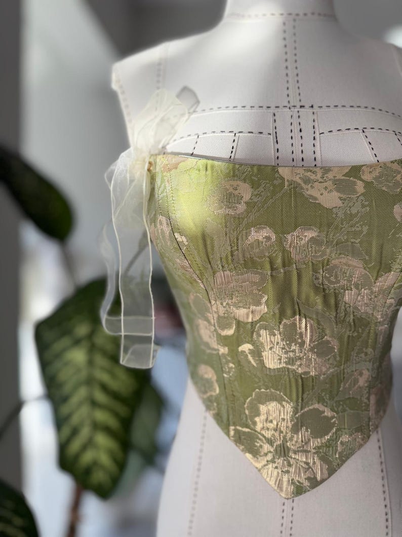 May include: A green and gold floral patterned corset top with a sweetheart neckline. The corset is displayed on a white dress form and features a delicate, sheer, white ribbon detail at the shoulder.