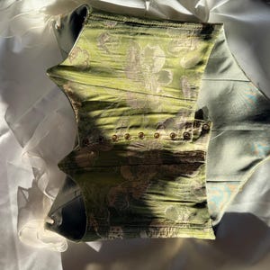 May include: A green corset with a floral pattern and gold-coloured eyelets. The corset is made of a textured fabric and has a structured design. The interior is a light blue colour.