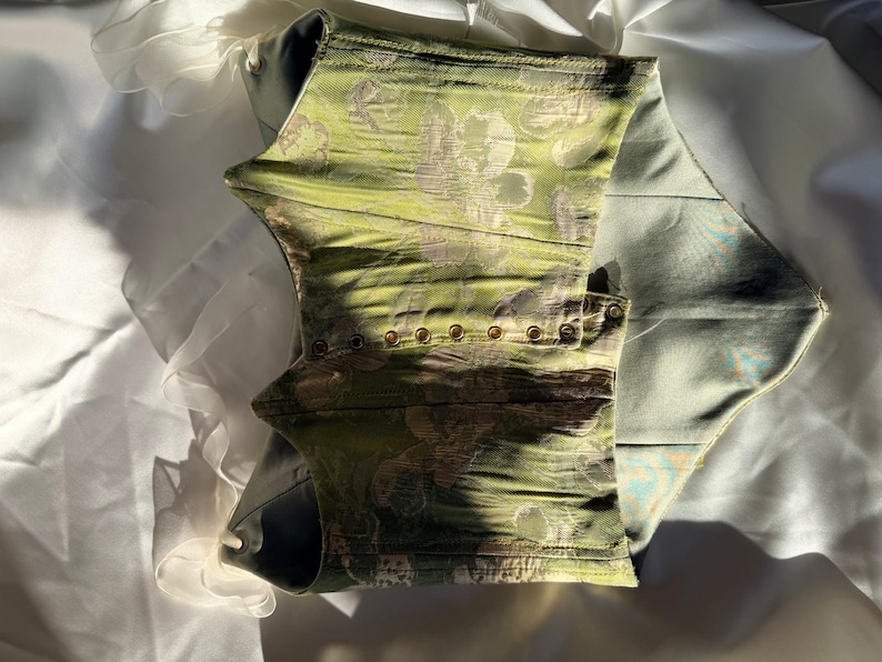 May include: A green corset with a floral pattern and gold-coloured eyelets. The corset is made of a textured fabric and has a structured design. The interior is a light blue colour.