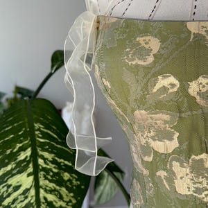 May include: A green and gold floral patterned corset with a white, sheer, ruffled ribbon detail. The corset is displayed on a white dress form. The background includes a large green plant.