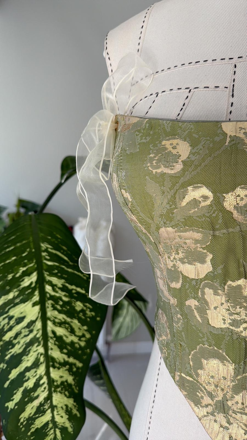 May include: A green and gold floral patterned corset with a white, sheer, ruffled ribbon detail. The corset is displayed on a white dress form. The background includes a large green plant.