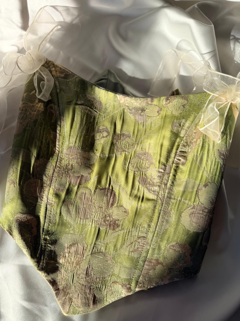 May include: A green corset featuring a floral design. The bodice is structured, and the shoulders are embellished with delicate, sheer, white bows. The fabric has a textured finish. The corset's design is elegant and vintage-inspired.