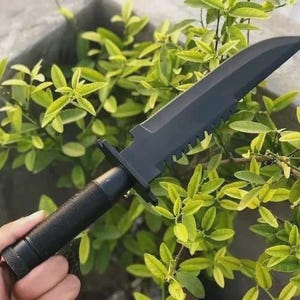 May include: A black tactical knife with a serrated edge and a textured handle. The knife is held against a backdrop of green foliage. The blade is approximately 8 inches long.