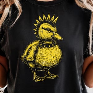 May include: Black t-shirt featuring a yellow punk rock duck graphic. The duck has a mohawk, a studded collar, and a detailed illustration style. The shirt is a casual wear item.