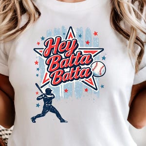 May include: White t-shirt featuring a baseball-themed graphic. The design includes a star, baseball, and a batter silhouette, with the text "Hey Batta Batta" in red and blue. The shirt is a classic crew neck.