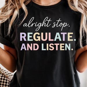 Puede incluir: Camiseta negra con la frase "alright stop. REGULATE. AND LISTEN." en blanco y colores pastel. La camiseta tiene mangas cortas y un corte relajado. El texto está en una fuente moderna y juguetona.
