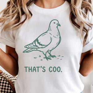 May include: White t-shirt featuring a green illustration of a pigeon and the phrase "THAT'S COO." The design is simple and clean, with the bird standing on a patch of grass. The shirt is a casual, everyday wear item.