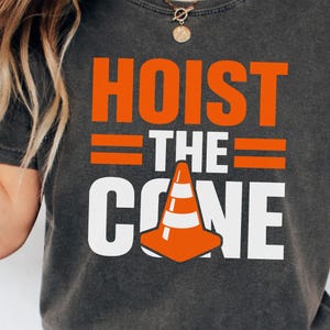 May include: Dark gray t-shirt with the words "HOIST THE CONE" in orange and white block letters. A graphic of an orange and white traffic cone is featured below the text. The shirt is a casual wear item.