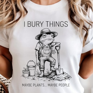 May include: White t-shirt with a humorous graphic of a frog in overalls and a hat, leaning on a shovel. The text reads "I BURY THINGS MAYBE PLANTS... MAYBE PEOPLE."