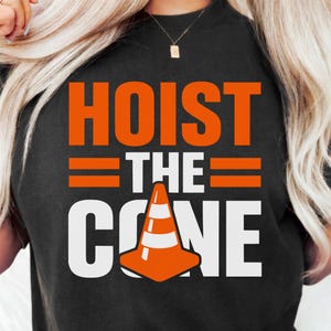 May include: Black t-shirt with the words "HOIST THE CONE" in orange and white block letters. The design includes an orange traffic cone graphic. The shirt is a casual style.