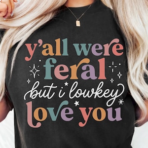 May include: Black t-shirt with the text "y'all were feral but i lowkey love you" in a colorful, retro font. The text is in shades of pink, teal, orange, and purple. The shirt has a crew neck and short sleeves.