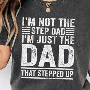 Può includere: T-shirt grigio scuro con la scritta bianca "I'M NOT THE STEP DAD I'M JUST THE DAD THAT STEPPED UP" invecchiata. La maglietta ha un girocollo e maniche corte. La maglietta è realizzata in un materiale morbido e confortevole.