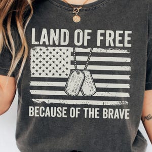 Op de afbeelding: Donkergrijze t-shirt met de tekst "LAND OF FREE BECAUSE OF THE BRAVE" boven een verweerde Amerikaanse vlag en hondenpenningen. Het shirt heeft een ronde hals en korte mouwen.