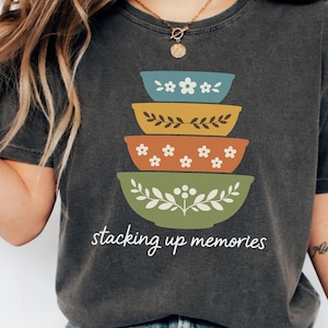 May include: Dark gray t-shirt featuring a stack of colorful bowls with floral and leaf designs. The bowls are in shades of blue, yellow, orange, and green. The text "stacking up memories" is printed below the bowls in a handwritten style.