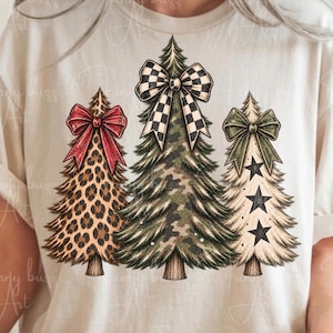 May include: A cream-coloured t-shirt featuring a graphic of three stylised Christmas trees. The left tree has a leopard print pattern and a red bow, the centre tree has a camouflage pattern and a black and white chequered bow, and the right tree has a cream base with black stars and a green bow.