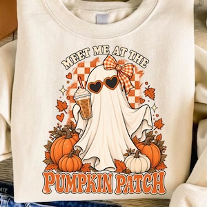 Pode incluir: Uma camisola de gola redonda cor de creme com um gráfico de um fantasma a usar óculos de sol em forma de coração e um laço xadrez laranja. O fantasma segura um café gelado, rodeado por abóboras cor de laranja, folhas de outono e o texto.