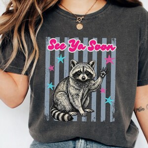 Peut inclure: T-shirt gris fonc&eacute; avec une illustration de raton laveur qui salue, avec le texte "See Ya Soon" en rose. Le motif comprend des &eacute;toiles bleues et roses et des rayures verticales grises. V&ecirc;tement d&eacute;contract&eacute;.