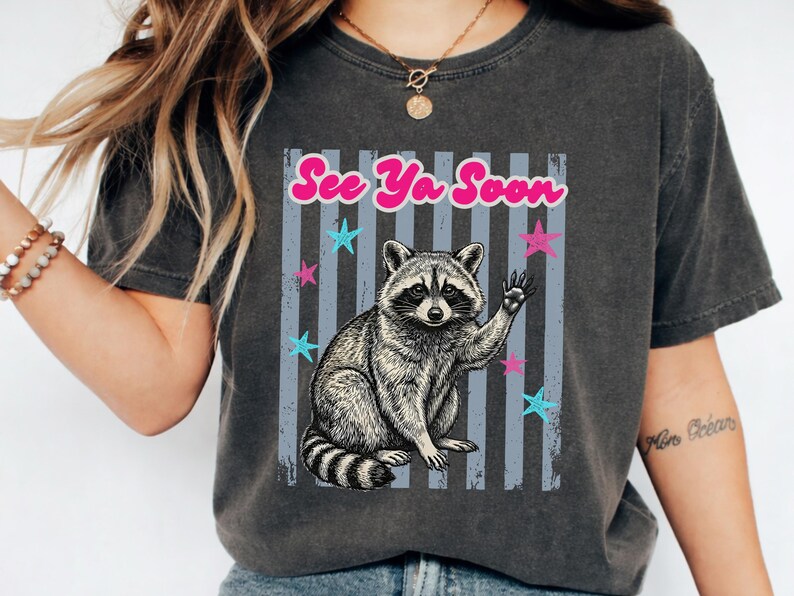 Peut inclure: T-shirt gris fonc&eacute; avec une illustration de raton laveur qui salue, avec le texte "See Ya Soon" en rose. Le motif comprend des &eacute;toiles bleues et roses et des rayures verticales grises. V&ecirc;tement d&eacute;contract&eacute;.