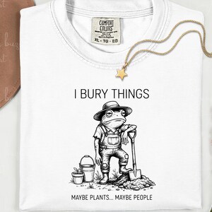 May include: White t-shirt with a graphic of a frog wearing overalls and a hat, holding a shovel. Text reads "I BURY THINGS MAYBE PLANTS... MAYBE PEOPLE". A gold star necklace and a brown hat are also visible.