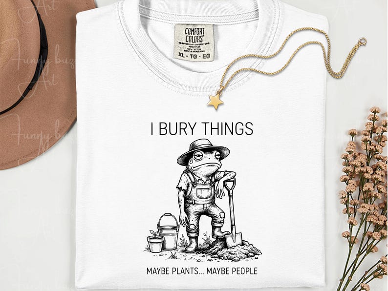 May include: White t-shirt with a graphic of a frog wearing overalls and a hat, holding a shovel. Text reads "I BURY THINGS MAYBE PLANTS... MAYBE PEOPLE". A gold star necklace and a brown hat are also visible.