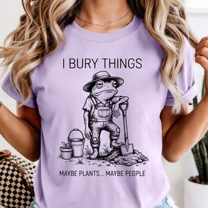 May include: A light purple t-shirt featuring a cartoon frog in overalls and a hat, leaning on a shovel. The text "I BURY THINGS" is above the frog, and "MAYBE PLANTS... MAYBE PEOPLE" is below.