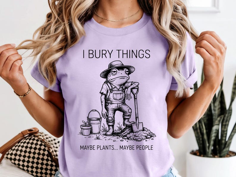 May include: A light purple t-shirt featuring a cartoon frog in overalls and a hat, leaning on a shovel. The text "I BURY THINGS" is above the frog, and "MAYBE PLANTS... MAYBE PEOPLE" is below.