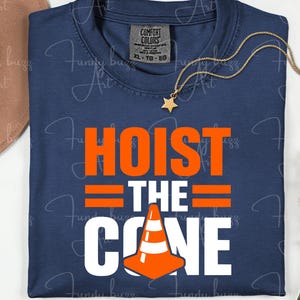 May include: Navy blue t-shirt with the words "HOIST THE CONE" in orange and white. The design includes an orange traffic cone graphic. A gold necklace with a star pendant and a brown hat are also visible.