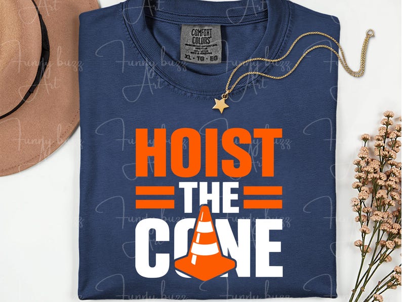 May include: Navy blue t-shirt with the words "HOIST THE CONE" in orange and white. The design includes an orange traffic cone graphic. A gold necklace with a star pendant and a brown hat are also visible.