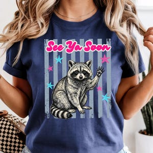 Peut inclure: T-shirt bleu marine avec un dessin de raton laveur qui salue. Le texte "See Ya Soon" est au-dessus du raton laveur, avec des &eacute;toiles et des rayures verticales. Le motif est imprim&eacute; sur un t-shirt &agrave; manches courtes.