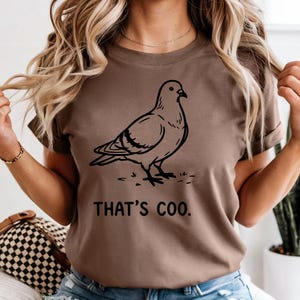 May include: Brown t-shirt featuring a black line drawing of a pigeon. The text "THAT'S COO." is printed below the bird. The shirt is a solid color and has short sleeves.