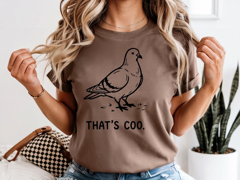 May include: Brown t-shirt featuring a black line drawing of a pigeon. The text "THAT'S COO." is printed below the bird. The shirt is a solid color and has short sleeves.