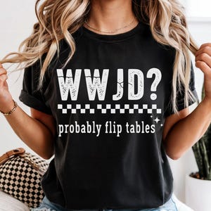 May include: Black t-shirt with the white text "WWJD?" above "probably flip tables." The design includes a checkered pattern and star accents. The shirt is a casual, comfortable style.