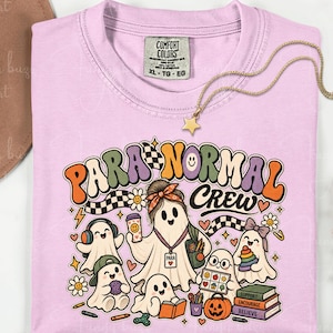 May include: A light pink crewneck t-shirt featuring a graphic print with the text "PARA NORMAL CREW". The design includes several cartoon ghosts, a pumpkin, books, and various small icons in orange, purple, black, and white colors.
