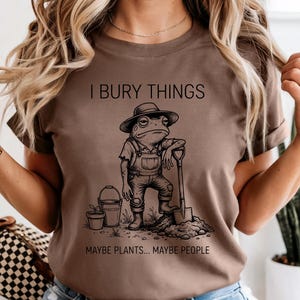 May include: Brown t-shirt with the text "I BURY THINGS MAYBE PLANTS... MAYBE PEOPLE." The graphic is a cartoon frog wearing overalls and a hat, leaning on a shovel. A bucket and potted plant are also in the image.