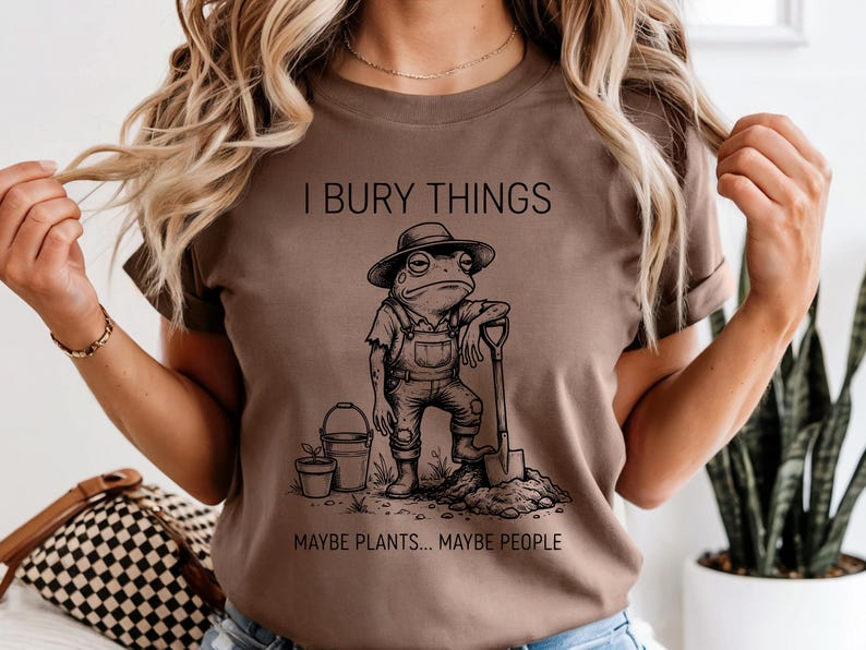May include: Brown t-shirt with the text "I BURY THINGS MAYBE PLANTS... MAYBE PEOPLE." The graphic is a cartoon frog wearing overalls and a hat, leaning on a shovel. A bucket and potted plant are also in the image.