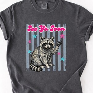 Peut inclure: T-shirt gris fonc&eacute; avec une illustration de raton laveur faisant un signe de la main et le texte "See Ya Soon". Le motif comprend des &eacute;toiles bleues et roses et des rayures verticales. Le t-shirt a des manches retrouss&eacute;es.