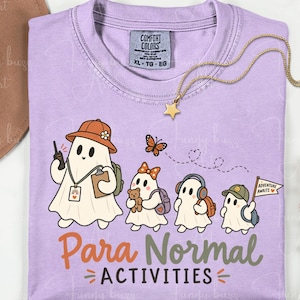 Peut inclure: Un t-shirt violet clair avec un motif représentant quatre fantômes prêts pour l'aventure. Ils portent des chapeaux, des sacs à dos et des accessoires comme un talkie-walkie, un ours en peluche et un drapeau. Le texte indique Para Normal ACTIVITIES.